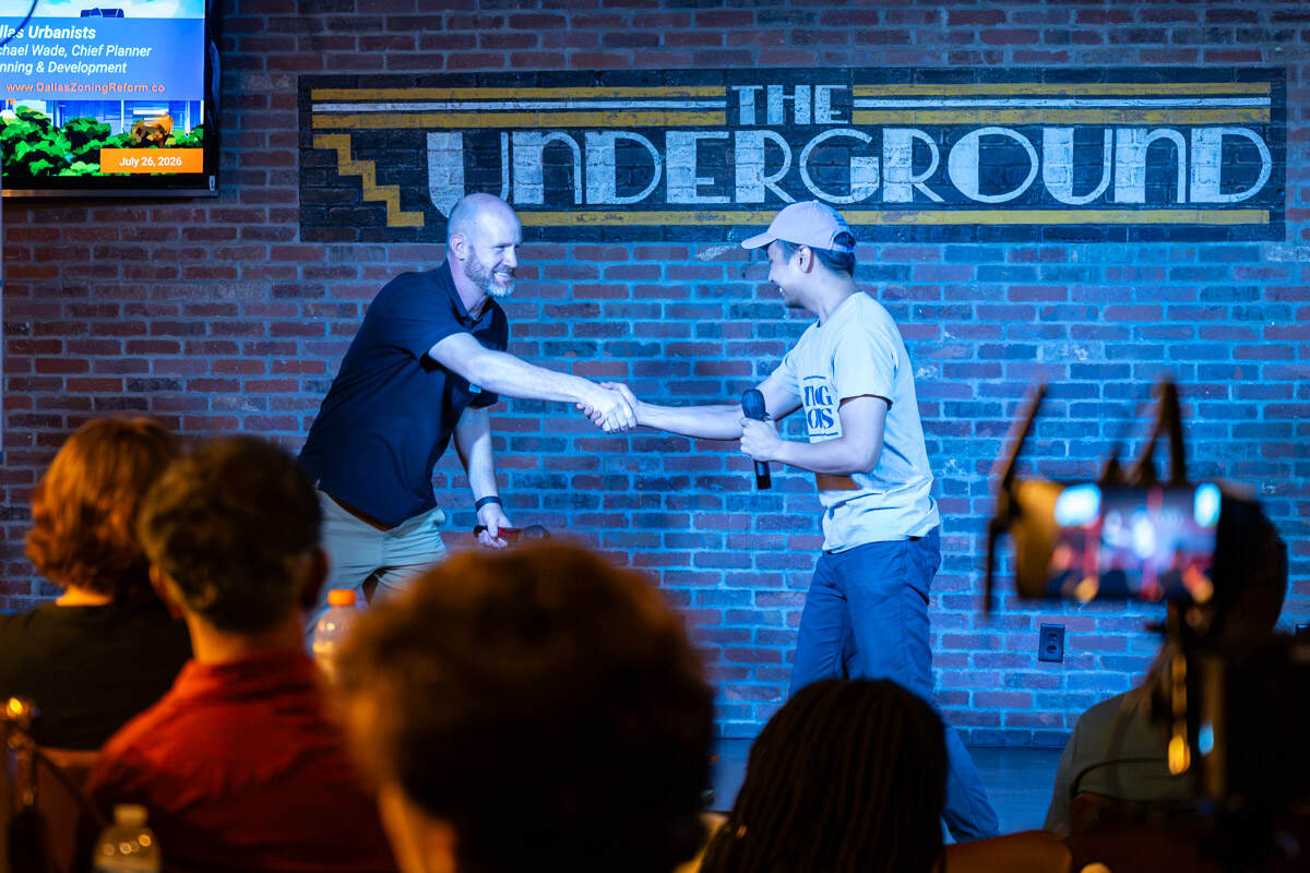 Two men shaking hands on stage in front of The Underground's painted brick-wall sign, with the audience in the foreground