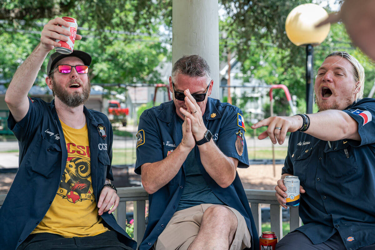 Three friends in matching navy work shirts laughing and toasting with beer cans in a park gazebo