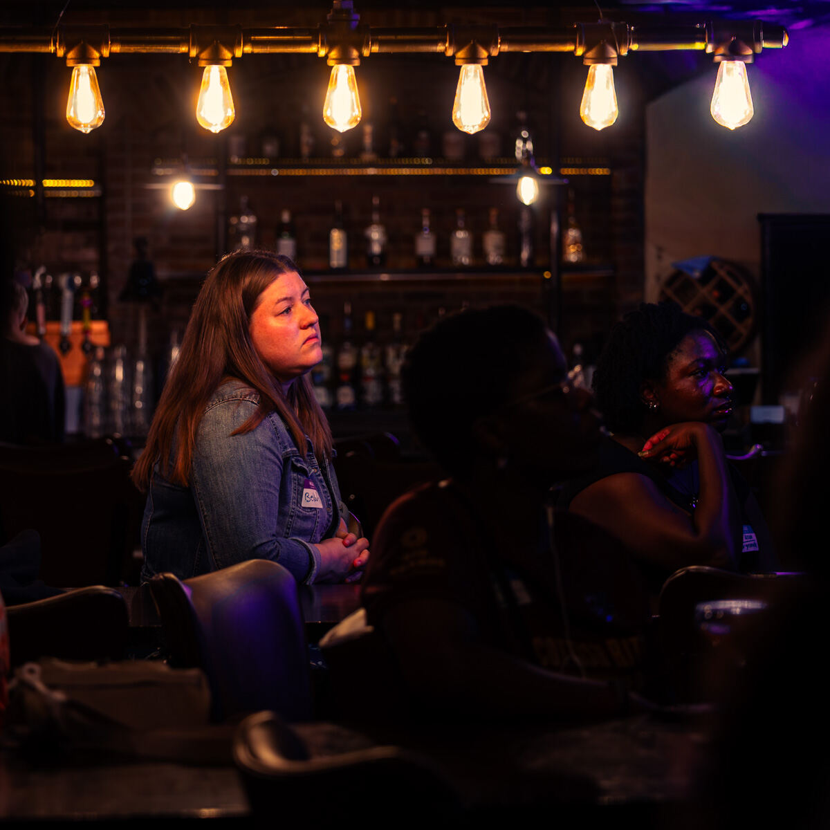 Woman in a denim jacket listening intently beneath a row of glowing Edison bulbs in a dim bar