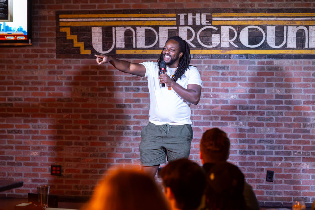 Performer with a microphone pointing out at the crowd on stage in front of The Underground's brick-wall sign