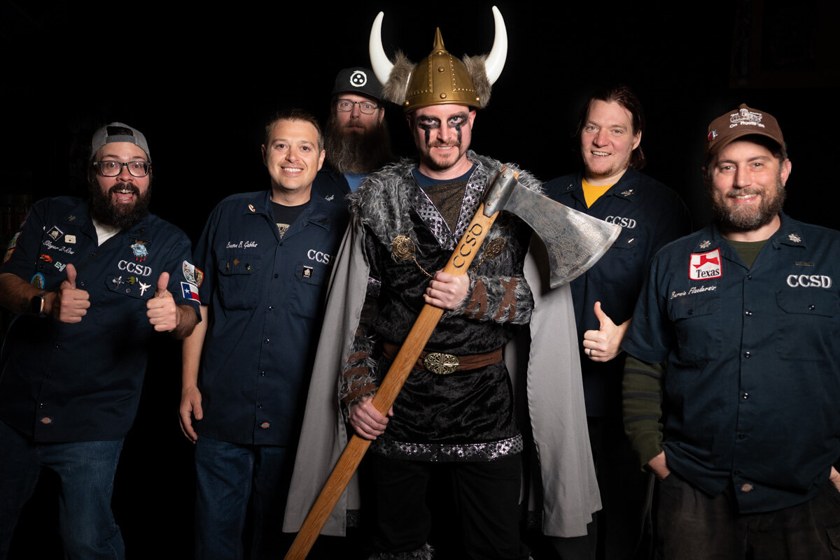 Group in matching navy work shirts giving thumbs up around a friend in a Viking costume holding an axe