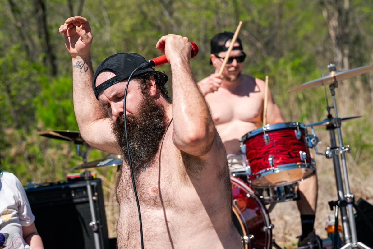 Bearded, shirtless singer with a red microphone performing outdoors as the drummer plays behind him