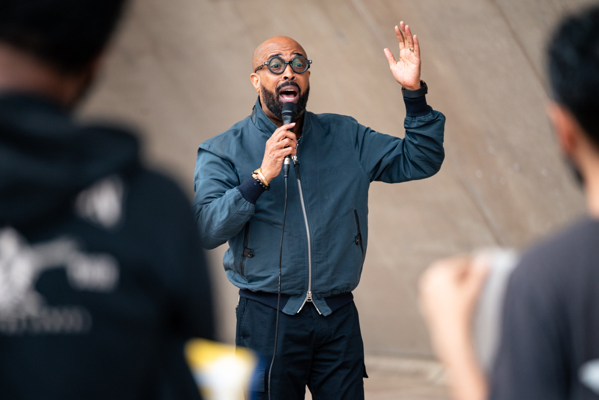Speaker in round glasses and a blue jacket addressing a crowd with one hand raised