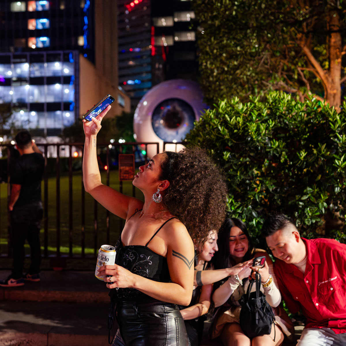 Woman holding up her phone at night beside the giant eyeball sculpture in downtown Dallas as friends look at a phone nearby