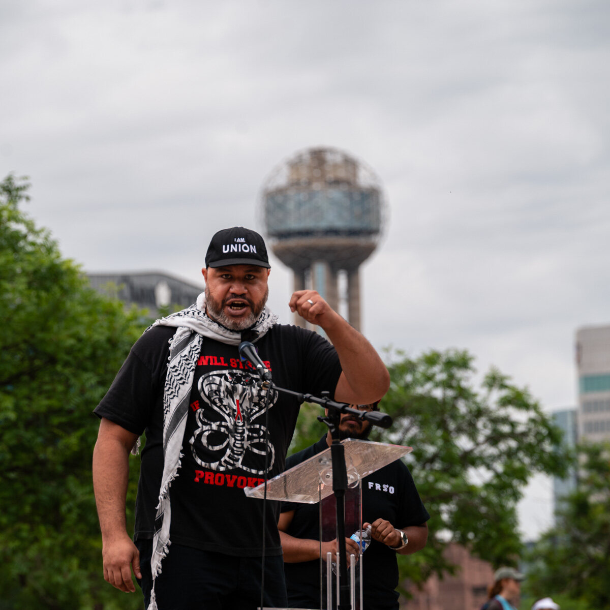Speaker in an I Am Union cap addressing a rally from a podium with Reunion Tower behind him